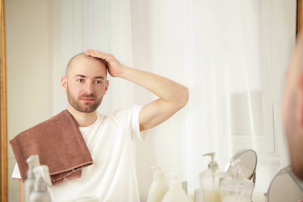 Young handsome bald man looking at mirror and touching his head